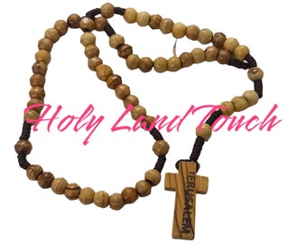 Può includere: Un rosario con perline in legno d'ulivo e un ciondolo a forma di croce in legno. La croce è incisa con la parola "Gerusalemme". Il rosario è infilato su un cordoncino marrone scuro. Il testo "Holy Land Touch" è nell'immagine.