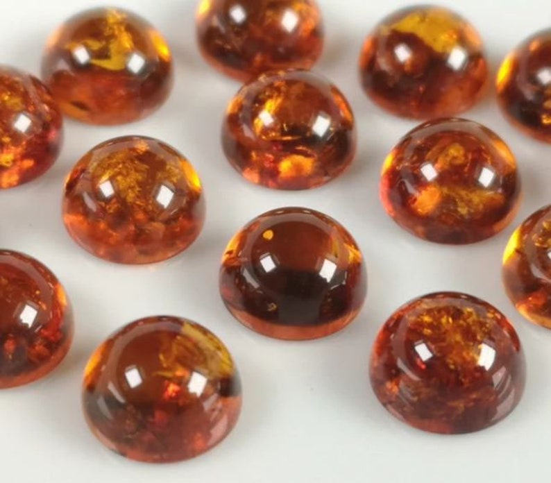 May include: A collection of round, amber-colored cabochons. The cabochons are smooth and have a glossy finish. They are arranged in a random pattern on a white surface.