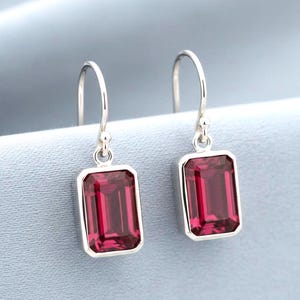 Pode incluir: Um par de brincos pendentes de prata com pedras preciosas vermelho-escuras de corte esmeralda retangular, engastadas em molduras de prata polida. Os brincos pendem de ganchos de prata simples, exibindo um design simétrico e limpo.