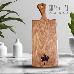 May include: A wooden chopping board with a maple leaf cutout. The board has a natural wood finish and a handle. The text "GRAY LEAF DESIGN CO." is printed on a white background.
