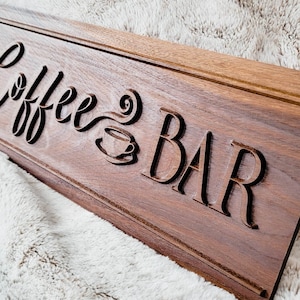 Peut inclure: Une enseigne en bois avec les mots "Coffee Bar" gravés dessus. L'enseigne est brune et a une finition en bois naturel. Les mots sont dans une police de caractères stylisée et il y a une petite image d'une tasse de café avec de la vapeur qui s'en échappe.