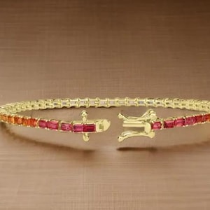 May include: A gold-toned bracelet with a gradient of orange to red gemstones. The bracelet is open, showcasing the clasp and the arrangement of the rectangular-cut stones. The background is a warm, brown tone.