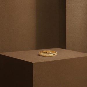Könnte beinhalten: Ein goldener Ring mit einem eingravierten Blumenmuster. Der Ring wird auf einer braunen Oberfläche präsentiert. Der Ring ist im Bandstil gehalten und hat ein detailliertes Design rund um den gesamten Umfang. Der Ring besteht aus glänzendem, gelbem Gold.