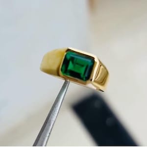 Könnte beinhalten: Ein goldener Ring mit einem rechteckigen, smaragdgeschliffenen grünen Edelstein. Der Ring hat ein dickes, poliertes Goldband. Der Edelstein ist in einer Lünettenfassung gefasst, was einen eleganten Look erzeugt. Der Ring wird von einer Pinzette gehalten.