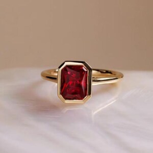 Könnte beinhalten: Ein goldener Ring mit einem rechteckigen, tiefroten Edelstein. Der Edelstein ist in einer Lünettenfassung gefasst, und das Band ist aus poliertem Gold. Der Ring wird auf einer weißen Oberfläche präsentiert.