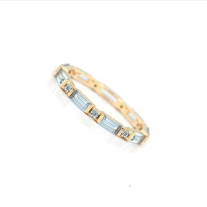Könnte beinhalten: Ein goldener Bandring mit einem abwechselnden Muster aus hellblauen Baguette-Schliff-Steinen und kleinen runden, klaren Steinen in einer durchgehenden Reihe. Das Metall hat eine polierte Oberfläche und ein schlankes, gleichmäßiges Profil.
