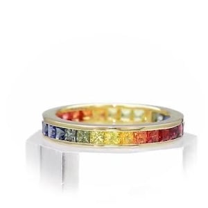 Könnte beinhalten: Ein goldener Bandring mit einer durchgehenden Reihe quadratisch geschliffener Edelsteine in Regenbogenfarben. Der Ring zeigt rote, orange, gelbe, grüne, blaue und violette Farbtöne. Er wird auf einem klaren, rechteckigen Ständer vor weißem Hintergrund präsentiert.
