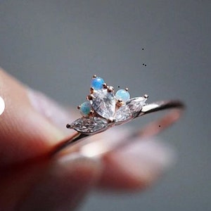 Könnte beinhalten: Ein zarter Ring aus Roségold mit einem floralen Design, das einen großen, klaren Mittel-Stein und kleinere blaue Steine aufweist.