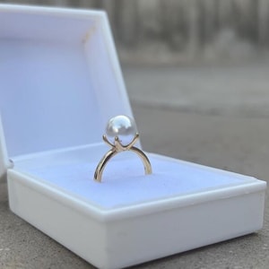 May include: A gold ring featuring a large, white pearl, presented in a four-prong setting. The ring is showcased within an open, white jewellery box with a soft, white interior. The background is blurred, implying an outdoor location.