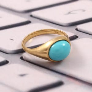 Könnte beinhalten: Ein goldfarbener Ring mit einem glatten, ovalen türkisfarbenen Stein. Der Ring hat ein schlichtes, klassisches Design mit poliertem Finish. Der Ring liegt auf einer weißen Oberfläche.