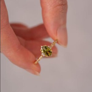 Könnte beinhalten: Ein goldener Ring mit einem ovalen, olivgrünen Edelstein, umgeben von kleinen, klaren Akzentsteinen. Das Band des Rings hat ein gedrehtes Design. Der Ring wird von einer Hand vor einem weichen, weißen Hintergrund gehalten.