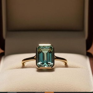 Könnte beinhalten: Ein goldener Ring mit einem rechteckigen, teal-farbenen Edelstein, gefasst in einem goldenen Band. Der Ring wird in einer offenen, braunen Schmuckschatulle mit cremefarbenem Innenraum präsentiert. Das Design des Rings ist schlicht und elegant.
