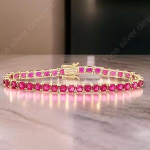 May include: A gold-colored bracelet with a series of square, vibrant red gemstones. The bracelet is clasped and rests on a light-colored surface, showcasing the jewelry's design and color.