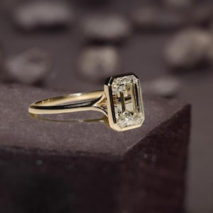 Könnte beinhalten: Ein goldener Ring mit einem Diamanten im Smaragdschliff. Der Diamant ist in einer rechteckigen Lünette gefasst, und das Band hat ein geteiltes Schaftdesign. Der Ring wird auf einer dunkelbraunen Oberfläche präsentiert.