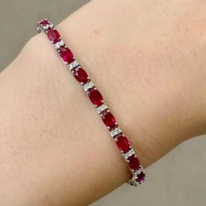Op de afbeelding: Een armband met ovale, dieprode edelstenen afgewisseld met kleine, heldere edelstenen. De armband is gezet in een zilverkleurig metaal en wordt getoond op een lichte huidskleur.
