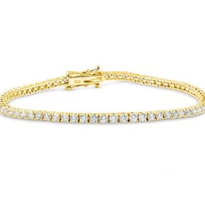 May include: A gold tennis bracelet featuring a line of small, clear gemstones. The bracelet has a secure clasp. The jewellery is set against a white background. A classic piece of jewellery.