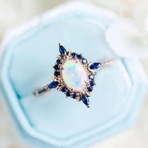 Puede incluir: Un anillo de oro rosa con una piedra central ovalada de ópalo iridiscente, rodeada de pequeñas piedras preciosas de color azul oscuro. El anillo está en una caja azul claro.