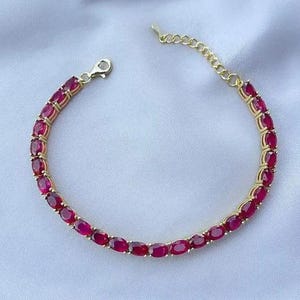 Puede incluir: Un brazalete dorado con piedras preciosas rojas oscuras de corte ovalado. El brazalete tiene una cadena delicada y un cierre. Las piedras están engastadas juntas, creando una línea continua de color.