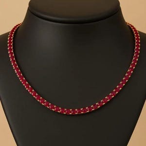 Op de afbeelding: Een goudkleurige ketting met een doorlopende rij helderrode edelstenen. De ketting wordt getoond op een zwarte mannequin, die het ontwerp en de kleur van het sieraad benadrukt. De edelstenen zijn dicht op elkaar geplaatst.