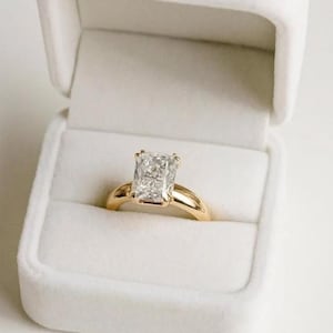 Puede incluir: Un anillo de compromiso con diamante de talla radiante y banda dorada descansa en un joyero de terciopelo blanco. El diamante es rectangular y brillante. La banda dorada tiene un acabado liso y pulido, creando un aspecto clásico.