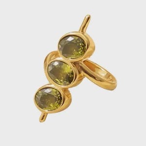 Könnte beinhalten: Ein goldfarbener Ring mit drei ovalen, olivgrünen Edelsteinen. Die Edelsteine sind in einzelnen Fassungen gefasst und vertikal angeordnet, mit einem kleinen Stab, der sich von oben und unten erstreckt. Der Ring hat ein schlichtes Band.