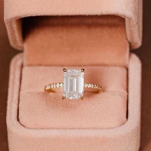 Puede incluir: Un anillo de oro con una gran gema clara rectangular como pieza central. La banda también es dorada y tiene gemas claras más pequeñas engastadas a los lados. El anillo se muestra dentro de un joyero de terciopelo rosa.