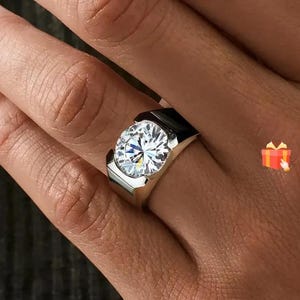 May include: A silver ring featuring a large, round, clear gemstone. The ring has a modern design with a thick band and a bezel setting. The gemstone sparkles brightly, reflecting light. The ring is a piece of jewellery.