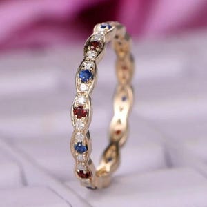 Puede incluir: Un anillo dorado con piedras preciosas rojas, azules y blancas alternas. El anillo presenta un patrón repetitivo de engastes ovalados, cada uno con una piedra preciosa. Las piedras son pequeñas y espaciadas.