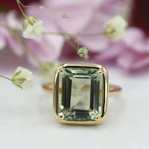 Könnte beinhalten: Ein goldener Ring mit einem großen, rechteckigen, hellgrünen Edelstein. Der Edelstein ist in einer goldenen Lünettenfassung gefasst. Der Ring wird auf einer weißen Oberfläche mit zarten rosa und weißen Blumen im Hintergrund gezeigt.