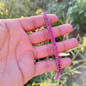 Könnte beinhalten: Ein silbernes Armband mit einer Reihe rechteckiger, leuchtend rosa Edelsteine. Das Armband wird in einer Hand gehalten, vor einem verschwommenen Hintergrund aus grünen Blättern und gelben Blüten. Die Edelsteine sind eng aneinandergereiht.
