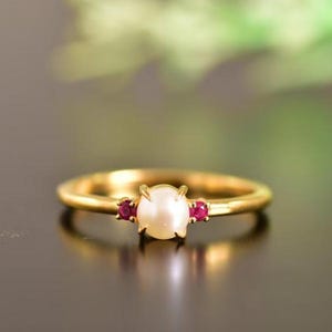 Könnte beinhalten: Ein goldener Ring mit einer zentralen Perle und zwei roten Edelsteinen. Das schlichte Design des Rings ist klassisch und elegant. Die Edelsteine sind beidseitig der Perle angeordnet.