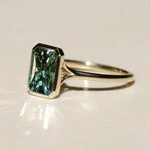 Könnte beinhalten: Ein goldener Ring mit einem rechteckigen, smaragdgeschliffenen, teal-grünen Edelstein. Der Edelstein ist in einer goldenen Lünettenfassung gefasst, und das Band ist aus poliertem Gold. Der Ring wird auf einer hellen Oberfläche gezeigt.