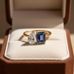 Könnte beinhalten: Ein goldener Ring mit zwei Edelsteinen: einem birnenförmigen, klaren Stein und einem rechteckigen, tiefblauen Stein. Der Ring ist in einem goldenen Band gefasst und wird in einer braunen Schmuckschatulle präsentiert.
