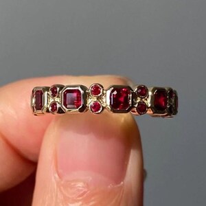 Peut inclure: Une bague en or ornée d'alternances de pierres rouges carrées et rondes. Le design de la bague présente un motif répétitif de pierres rouges vives serties dans un anneau en or poli, créant un bijou saisissant et élégant.