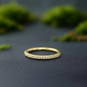 Könnte beinhalten: Ein goldener Bandring mit kleinen, klaren Steinen, die oben eingesetzt sind. Der Ring liegt auf einer dunklen Oberfläche mit verschwommenem grünem Laub im Hintergrund. Der Ring ist ein zartes Schmuckstück.