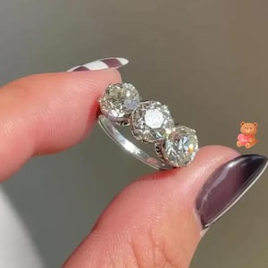 May include: A silver ring featuring three large diamonds. The ring has a classic design with three round-cut diamonds set in a row. The diamonds are clear and sparkling. The ring is held between fingers against a blurred background.