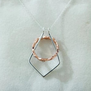 May include: A silver necklace featuring a geometric pendant. The pendant has a diamond-shaped frame with a rose gold ring suspended inside. The ring has a textured design. The necklace chain is also silver.