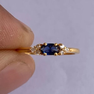Könnte beinhalten: Ein goldener Ring mit einem blauen ovalen Saphir und kleinen weißen Diamanten, die darum herum gesetzt sind.