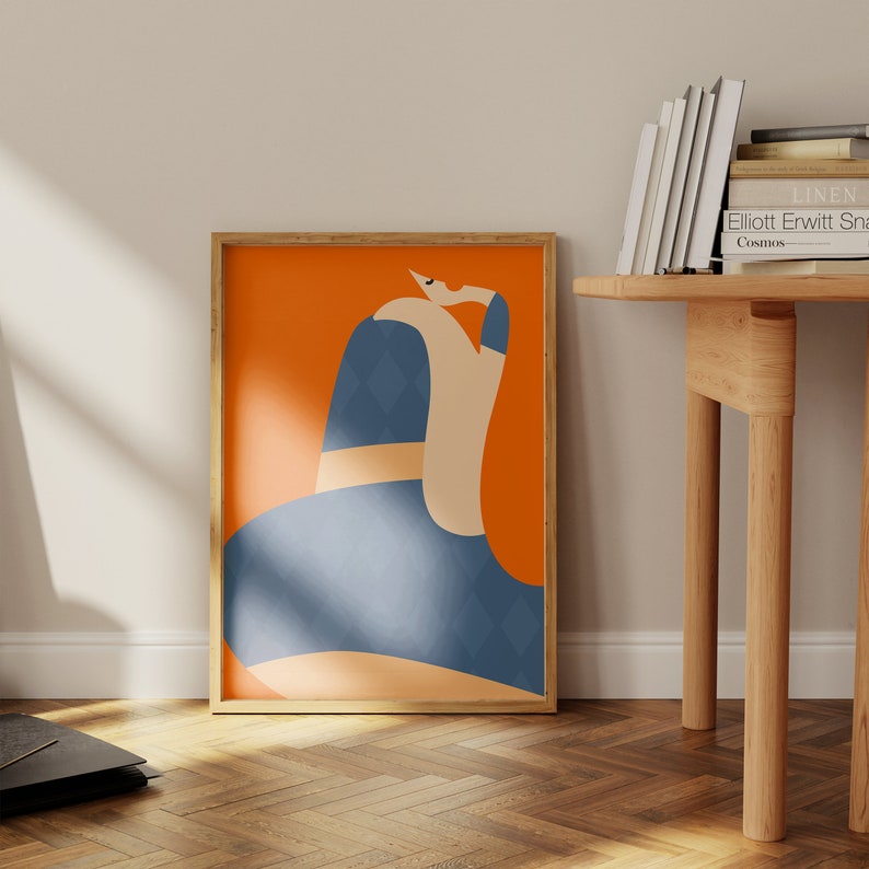 May include: A framed art print with an abstract illustration. The artwork shows a figure with a blue diamond-patterned form, set against an orange background. The print is encased in a natural wood frame, adding warmth to the design.