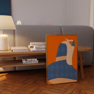 May include: A framed art print with an abstract design, set against an orange backdrop. The figure is rendered in blue, tan, and white. The artwork is displayed on a wooden floor, leaning against a gray sofa.