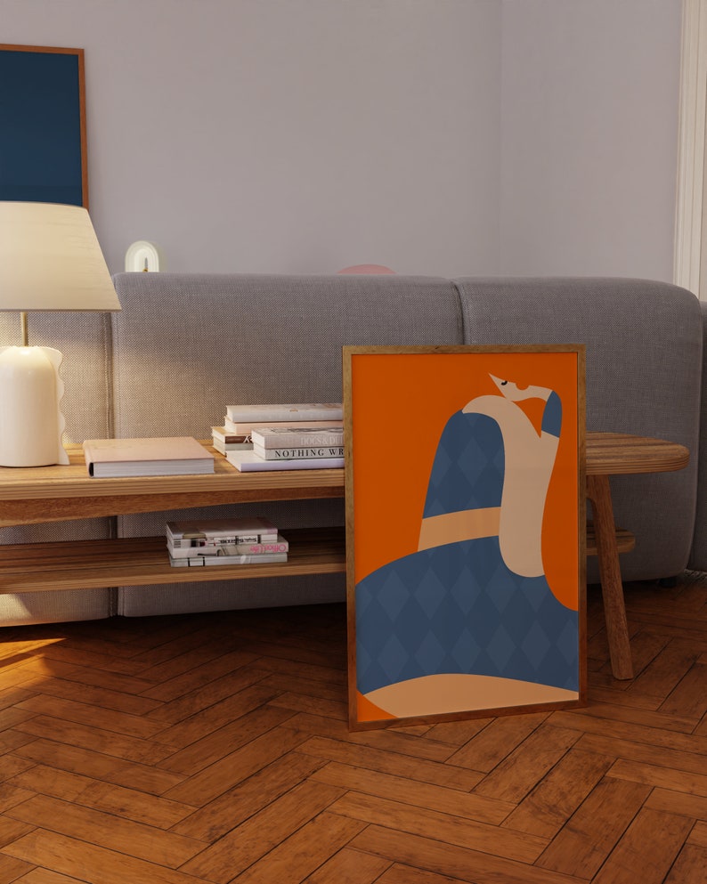 May include: A framed art print with an abstract design, set against an orange backdrop. The figure is rendered in blue, tan, and white. The artwork is displayed on a wooden floor, leaning against a gray sofa.