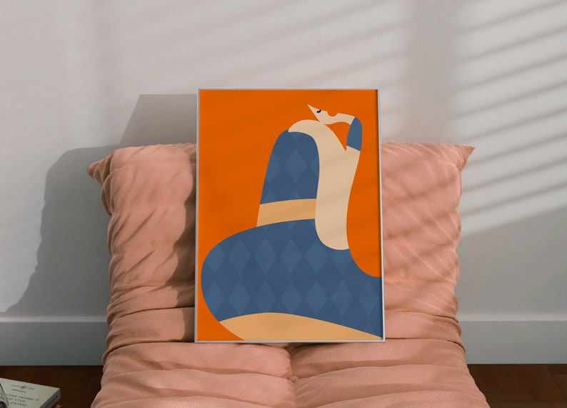 May include: An art print in a silver frame, depicting an abstract figure in a blue diamond-patterned dress against an orange backdrop. The artwork is displayed on a pink cushion, creating a modern aesthetic.