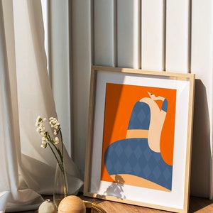 May include: An art print in a light wood frame, depicting an abstract figure with a blue diamond pattern on an orange background. The framed artwork leans against a white wall. A gold tray with decorative items and flowers sits on the floor.