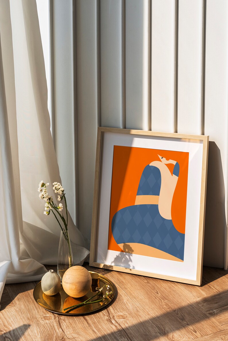 May include: An art print in a light wood frame, depicting an abstract figure with a blue diamond pattern on an orange background. The framed artwork leans against a white wall. A gold tray with decorative items and flowers sits on the floor.