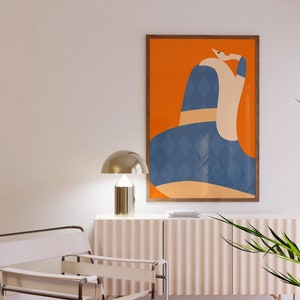 May include: An abstract art print in a wooden frame. The artwork features a figure with a blue diamond pattern, set against an orange background. A gold-coloured lamp sits on a white cabinet. A white chair with leather straps is in the foreground.