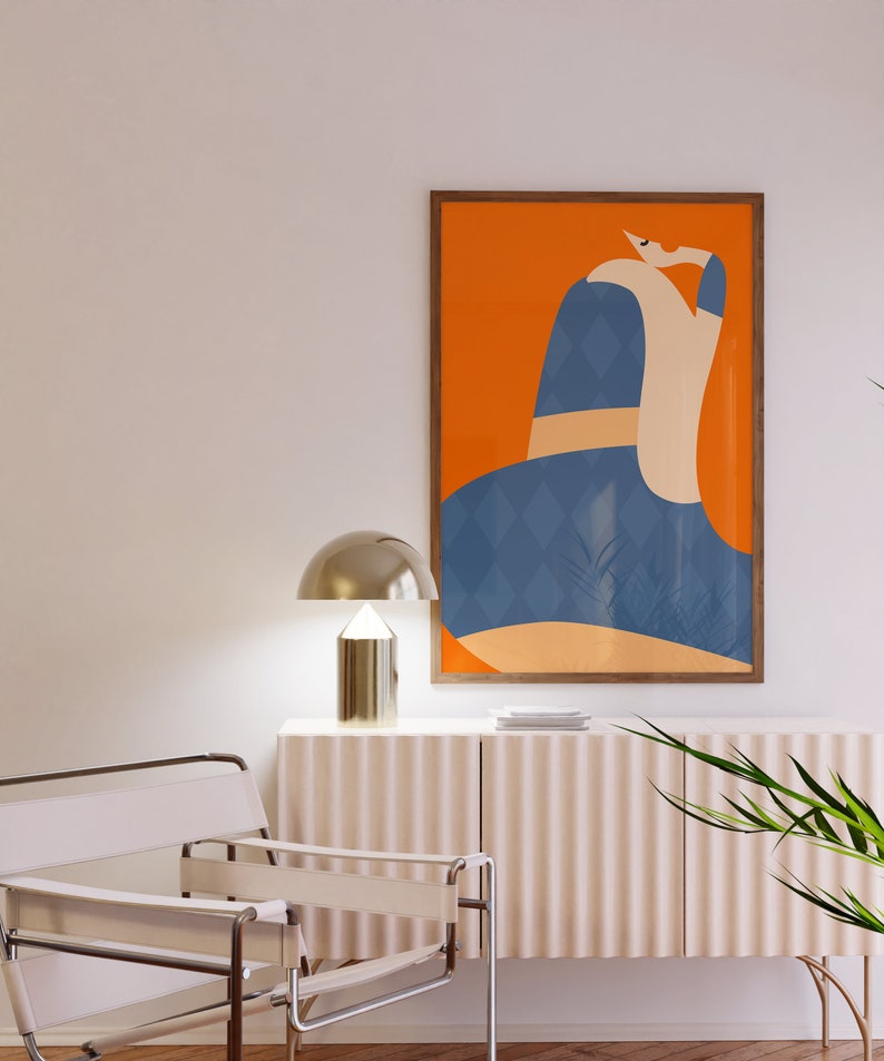 May include: An abstract art print in a wooden frame. The artwork features a figure with a blue diamond pattern, set against an orange background. A gold-coloured lamp sits on a white cabinet. A white chair with leather straps is in the foreground.