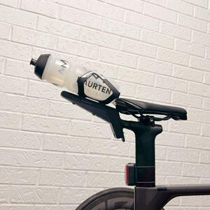 May include: A black bicycle saddle mounted on a seat post with a black bottle cage attached to the rear. A clear plastic water bottle with a black cap and the text MAURTEN printed in black is secured within the cage. A small red light is visible.