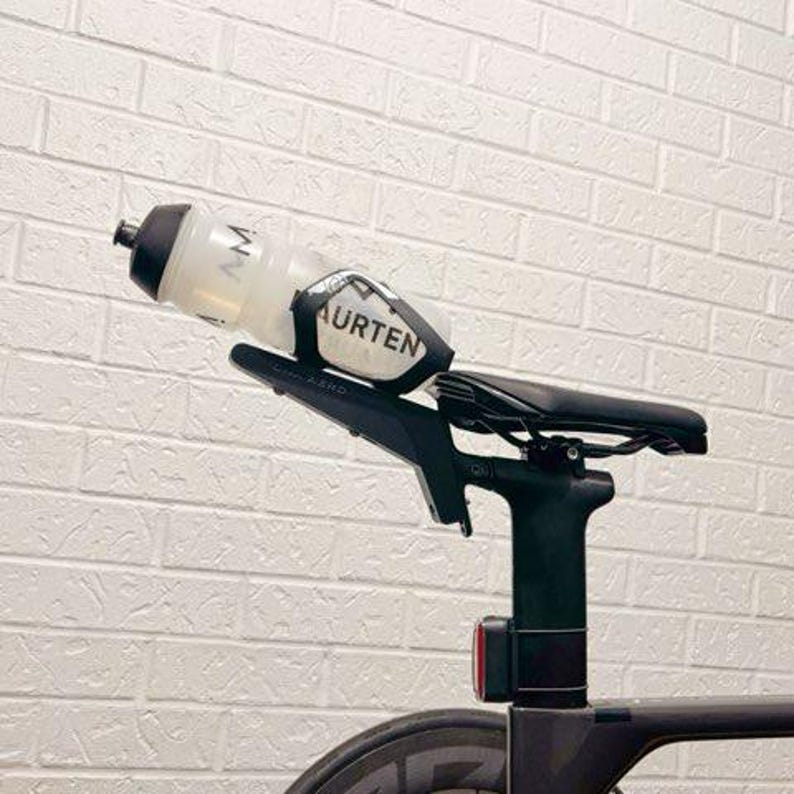 May include: A black bicycle saddle mounted on a seat post with a black bottle cage attached to the rear. A clear plastic water bottle with a black cap and the text MAURTEN printed in black is secured within the cage. A small red light is visible.