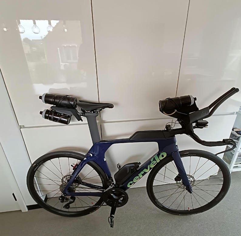May include: A dark blue Cerv&eacute;lo triathlon bicycle featuring a streamlined frame, aero handlebars with integrated water bottle mounts, two rear-mounted water bottles behind the seat, a frame-mounted storage box, and black disc brake wheels.