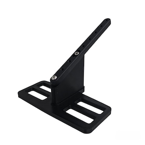 Peut inclure: Un support en métal noir avec une base rectangulaire comportant deux découpes rectangulaires. Un bras de support incliné est fixé à la base avec une série de trous percés sur toute sa longueur. Un deuxième bras plus court relie la base et le bras incliné.